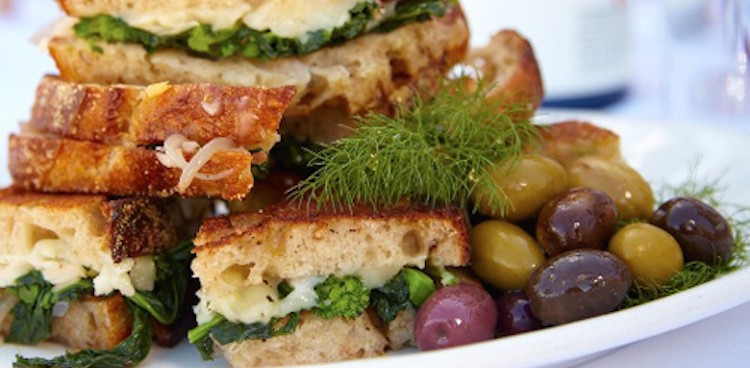 A stack of fontina and rapini grilled cheese sandwiches garnished with dill and served with a side of olives