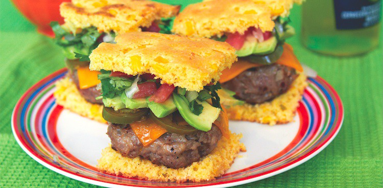 three sliders on cornbread topped with avocado, tomato, and cheddar and sandwiched between cornbread