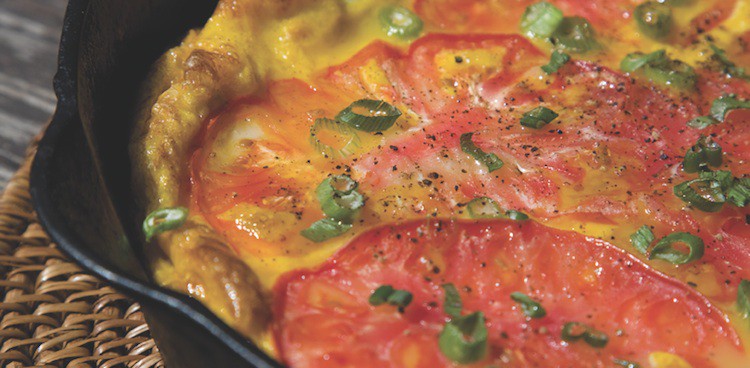 Frittata in a cast iron pan with golden cheese, red tomato slices, and sprinkled scallions