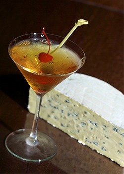 A Widow's Kiss Cocktail stands in a martini glass next to a half wedge of blue cheese.