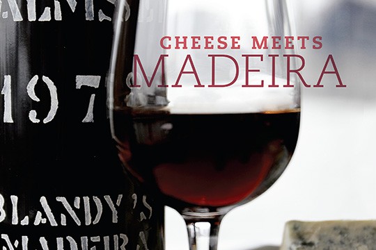 A glass of madeira stands next to a bottle and a hunk of crumbly cheese.