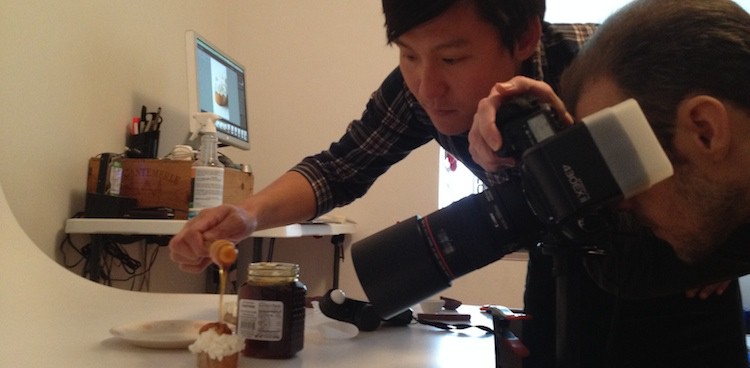 food stylist drips honey onto a piece of bread topped with ricotta cheese while a photographer stands read