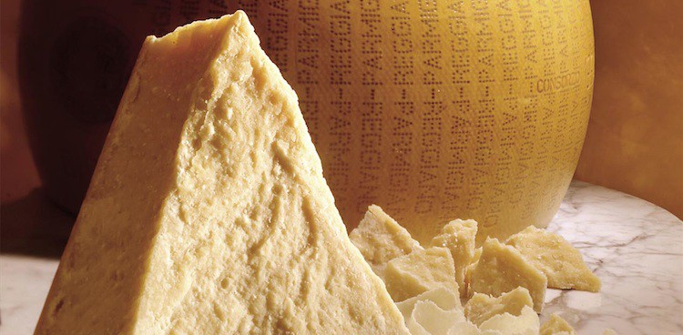 Wedge of Parmigiano Reggiano cheese in front of a parmesan wheel, crumbles, and shavings