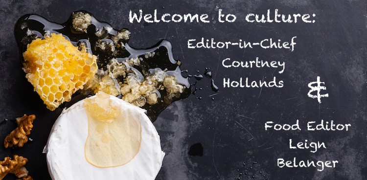 Cheese slate with bloomy rind cheese, honey, and nuts and the words "Welcome to culture editor-in-chief Courtney Hollands & food editor Leigh Belanger" written on it in chalk