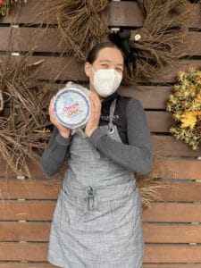 A woman wearing a mask holds a cheese wheel.