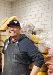 A man with glasses and a baseball hat poses among cheese with a white-tiled wall in the background.