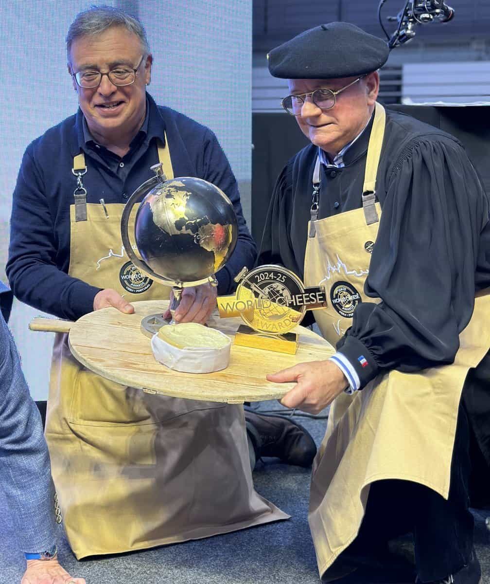 Two men hold a cheese on a large cheese board with a trophy.